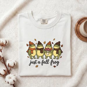 Könnte beinhalten: Ein weißes T-Shirt mit einer Grafik von vier grünen Fröschen, die Strickmützen und Schals tragen, wobei ein Frosch einen rot-weißen Pilz hält. Der Text "just a fall frog" ist in schwarzer Schrift unter den Fröschen gedruckt, umgeben von Herbstblättern.