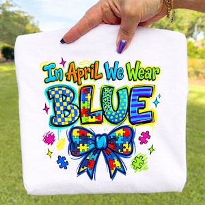 May include: White t-shirt with the text "In April We Wear BLUE" in colourful, stylised lettering. The word "BLUE" features various patterns, including leopard print and puzzle pieces. A bow tie made of puzzle pieces is at the bottom.