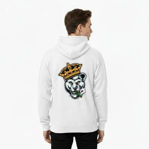 Op de afbeelding: Witte hoodie met een afbeelding van een gekroonde panter die een sigaret rookt. De panter draagt een gouden kroon en heeft een zwart-wit gezicht. De hoodie heeft een capuchon met trekkoord en lange mouwen.