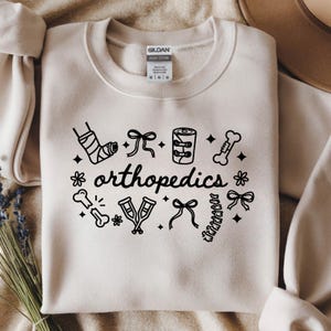 Op de afbeelding: Een crèmekleurige sweatshirt met het woord "orthopedie" in een zwarte, gestileerde lettertype. Het ontwerp bevat zwarte illustraties van een beengips, verbanden, botten en krukken. De sweatshirt heeft een ronde hals en lange mouwen.
