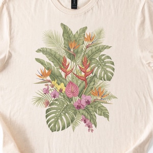 Könnte beinhalten: Ein naturfarbenes Kurzarm-T-Shirt mit einem zentralen Grafikdruck verschiedener tropischer Pflanzen und Blumen, darunter grüne Monstera-Blätter, orangefarbene Paradiesvogelblumen, rote Helikonien und kleine rosa Orchideen.