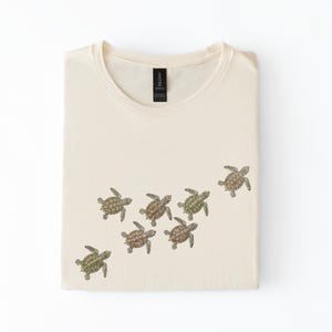 May include: Cream-coloured t-shirt with a design of nine small sea turtles in green, brown, and tan. The turtles are arranged diagonally across the shirt. The shirt is neatly folded.