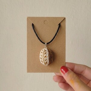 Peut inclure: Un pendentif en forme de mini miche de pain au levain sur un collier en cordon noir. Le pendentif présente une croûte texturée brun doré avec un motif de feuilles incisé sur le dessus. Il est fixé à une bélière argentée sur un support en carton.