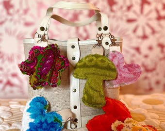 Könnte beinhalten: Eine kleine, cremefarbene Handtasche mit einem beigefarbenen Canvas-Körper und weißen Ledergriffen und -verzierungen. Befestigt sind mehrere bunte Stoff-Schlüsselanhänger in Form eines Herzens, eines Pilzes und einer Blume. Die Schlüsselanhänger sind in Rosa-, Grün-, Rot-, Blau- und gemusterten Stoffen gehalten.