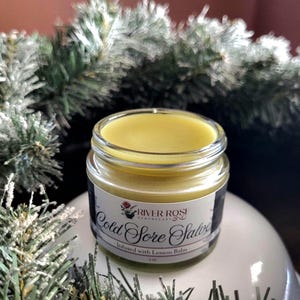 Puede incluir: Un pequeño frasco de vidrio lleno de ungüento amarillo. El frasco tiene una etiqueta blanca con texto negro que dice "River Rose Apothecary Cold Sore Salve Infused with Lemon Balm". El frasco está rodeado por un fondo borroso verde y blanco.