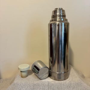 May include: A tall, stainless steel thermos with a matching cup and stopper. The thermos is cylindrical with a screw-on cap. The cup is a small, silver cylinder. The stopper is white and beige. The items are on a white towel.