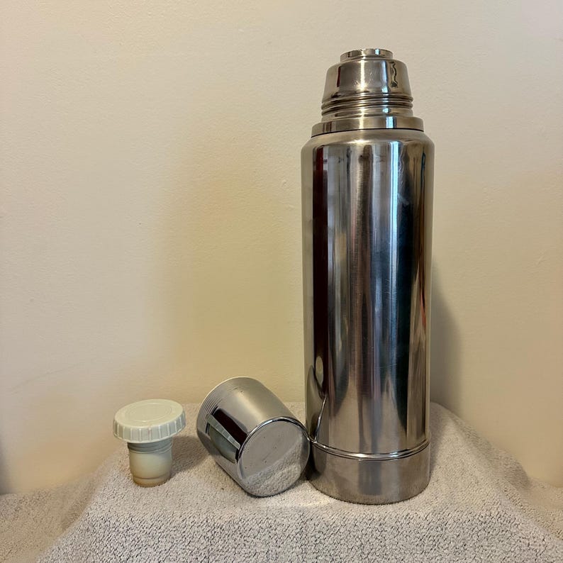 May include: A tall, stainless steel thermos with a matching cup and stopper. The thermos is cylindrical with a screw-on cap. The cup is a small, silver cylinder. The stopper is white and beige. The items are on a white towel.
