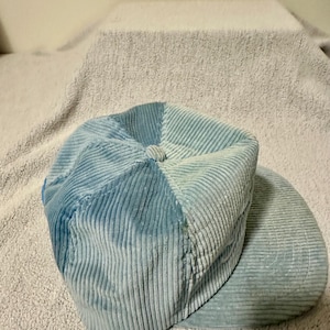 Op de afbeelding: Een lichtblauwe corduroy newsboy cap met een afgeronde kroon en een korte, stijve klep. De pet is gemaakt van een zachte, geribbelde stof en heeft een knoopdetail aan de bovenkant. De hoed is een stijlvol accessoire.