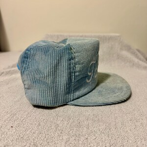 Op de afbeelding: Lichtblauwe corduroy baseballcap met een gebogen klep. De cap heeft witte geborduurde letters aan de zijkant. De hoed is gemaakt van een zachte, geribbelde stof.