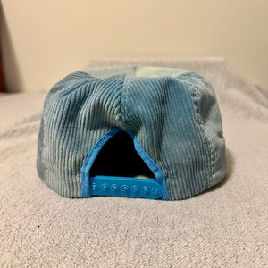 Op de afbeelding: Een lichtblauwe corduroy baseballcap met een snapbacksluiting. De pet heeft een zes-panelen ontwerp met een gebogen klep en een effen lichtblauwe kleur. De verstelbare band is ook lichtblauw.