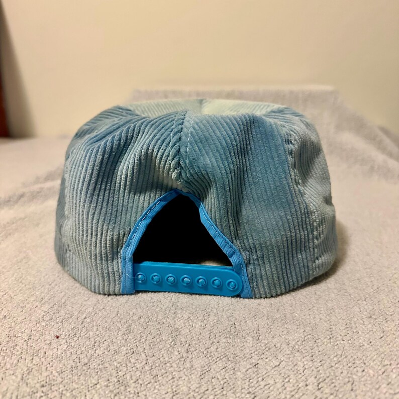 Op de afbeelding: Een lichtblauwe corduroy baseballcap met een snapbacksluiting. De pet heeft een zes-panelen ontwerp met een gebogen klep en een effen lichtblauwe kleur. De verstelbare band is ook lichtblauw.