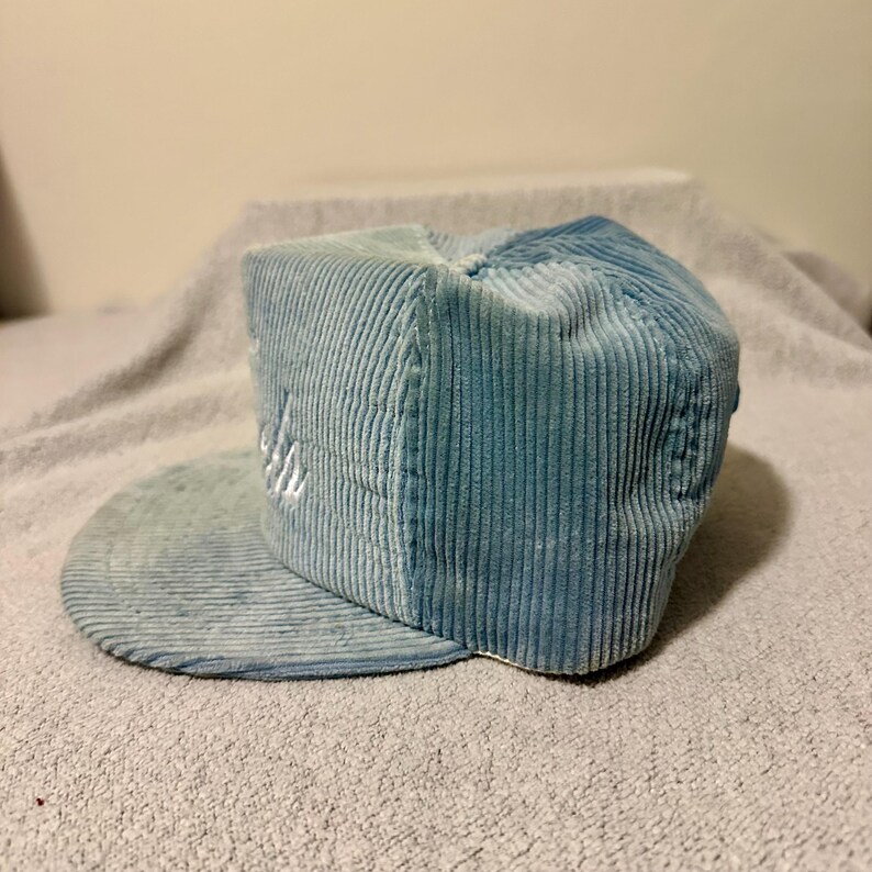 Op de afbeelding: Lichtblauwe corduroy baseballpet met een platte klep. De pet heeft een wit geborduurd ontwerp op de voorkant. De hoed is gemaakt van een zachte, geribbelde stof.