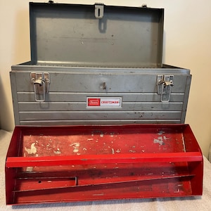 May include: An open, vintage Craftsman toolbox with a red tray and a gray metal exterior. The toolbox has a rectangular shape with a hinged lid and a latch closure. The Craftsman logo is visible on the front.