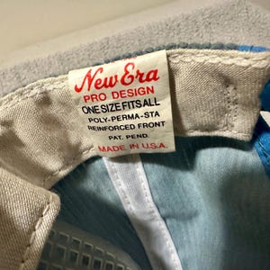 Op de afbeelding: Close-up van een New Era baseball cap tag. De tag is wit met rode tekst die luidt "New Era PRO DESIGN ONE SIZE FITS ALL POLY-PERMA-STA REINFORCED FRONT PAT. PEND. MADE IN U.S.A." De cap is lichtblauw en wit.