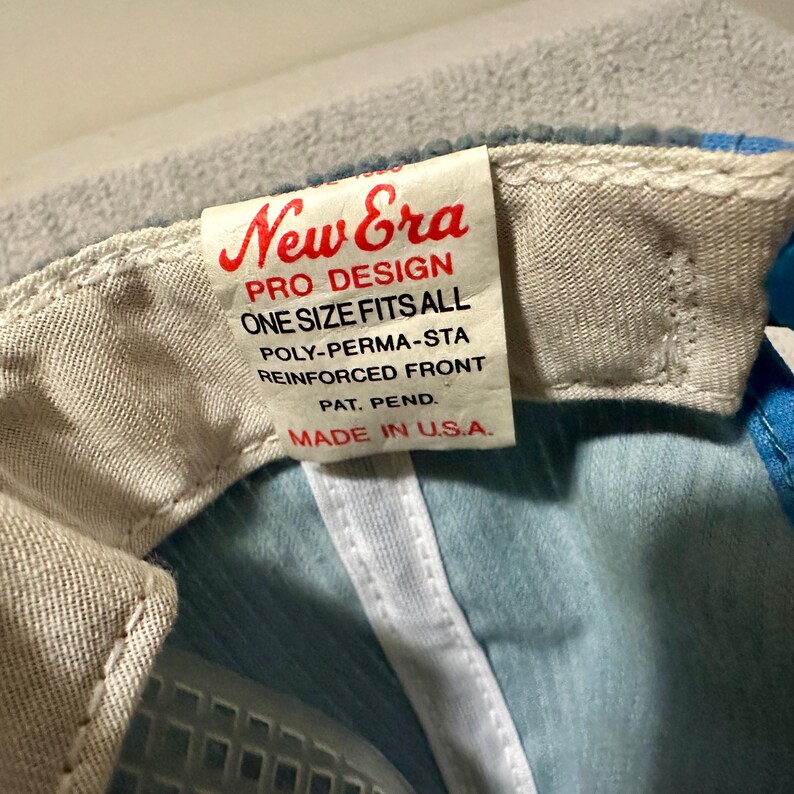 Op de afbeelding: Close-up van een New Era baseball cap tag. De tag is wit met rode tekst die luidt "New Era PRO DESIGN ONE SIZE FITS ALL POLY-PERMA-STA REINFORCED FRONT PAT. PEND. MADE IN U.S.A." De cap is lichtblauw en wit.