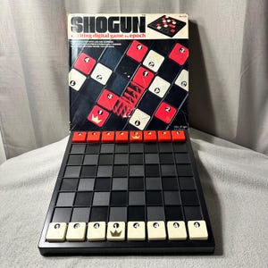 Puede incluir: Un juego de mesa Shogun vintage de epoch. El juego presenta un tablero negro y rojo con un patrón de cuadrícula. La caja muestra el nombre del juego y un ejemplo de diseño. El juego incluye piezas rojas y blancas con números y símbolos.