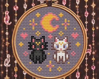 May include: A cross-stitch pattern in a round wooden hoop featuring a black cat and a white cat with crescent moon symbols on their foreheads, surrounded by a yellow crescent moon, pink hearts, and small yellow stars on a gray fabric.