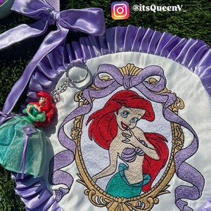Op de afbeelding: Geborduurde Ariel-patch met een paarse satijnen lintrand. De patch heeft een geborduurd beeld van Ariel uit De Kleine Zeemeermin, met een gouden frame en een paarse strik. Extra Ariel-thema accessoires zijn zichtbaar.