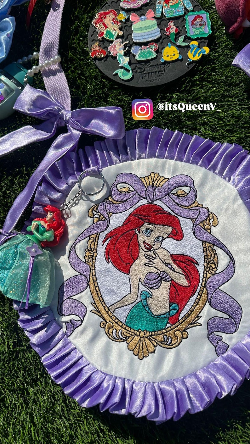 Op de afbeelding: Geborduurde Ariel-patch met een paarse satijnen lintrand. De patch heeft een geborduurd beeld van Ariel uit De Kleine Zeemeermin, met een gouden frame en een paarse strik. Extra Ariel-thema accessoires zijn zichtbaar.