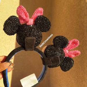May include: Black and pink Minnie Mouse ear headbands. The ears are covered in black rhinestones, and the bows are pink. The headband is black with a blue interior. The text "RnRcreations" is visible.