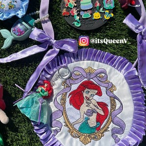 Op de afbeelding: Een verzameling items met het thema De Kleine Zeemeermin, waaronder een ronde, wit en paars geborduurde patch met Ariel's portret, een sleutelhanger, pluche speelgoed en emaille pins. De items zijn gerangschikt op een groene ondergrond.