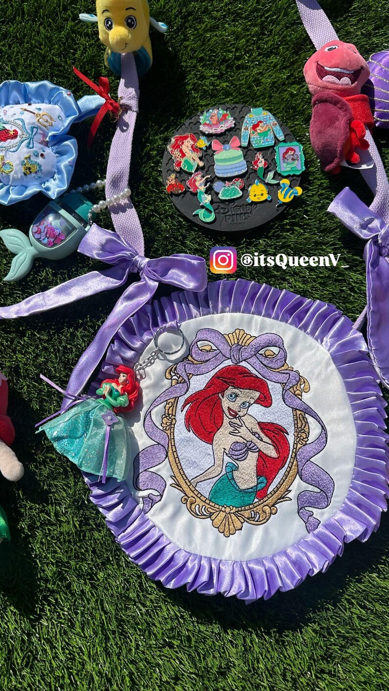 Op de afbeelding: Een verzameling items met het thema De Kleine Zeemeermin, waaronder een ronde, wit en paars geborduurde patch met Ariel's portret, een sleutelhanger, pluche speelgoed en emaille pins. De items zijn gerangschikt op een groene ondergrond.