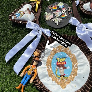 May include: A brown fabric strap featuring a round white satin display board with a gold embroidered Toy Story frame and Woody character. The board has a brown ruffled edge, white satin bows, and a hanging Woody figurine keychain attached.