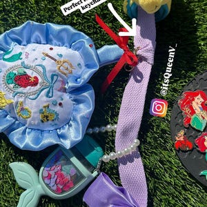 Op de afbeelding: Een verzameling accessoires met het thema De Kleine Zeemeermin, waaronder een pluche Botje, een blauw-wit geborduurd zakje, een handdesinfectiehouder in de vorm van een zeemeerminstaart en een paarse lanyard met een strik.