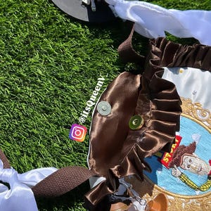 May include: A brown and white fabric bag featuring a ruffled brown satin trim, a circular embroidered patch of a cowboy character, and two attached toy figures. The bag includes white ribbon ties, brown woven straps, and a metal snap closure.
