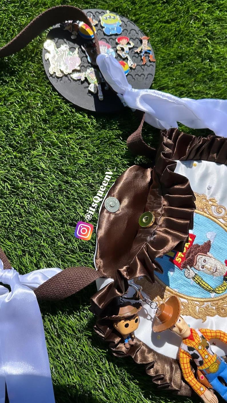 May include: A brown and white fabric bag featuring a ruffled brown satin trim, a circular embroidered patch of a cowboy character, and two attached toy figures. The bag includes white ribbon ties, brown woven straps, and a metal snap closure.