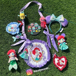 Op de afbeelding: Een verzameling Ariel-thema accessoires, waaronder pluche poppen, een hoofdband met een strik, een ronde tas en diverse bedels. De items tonen de afbeelding van de Kleine Zeemeermin en zijn in tinten paars, rood en blauwgroen, tegen een groene achtergrond.