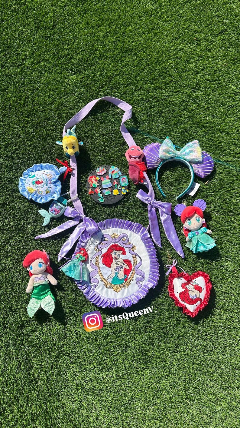 Op de afbeelding: Een verzameling Ariel-thema accessoires, waaronder pluche poppen, een hoofdband met een strik, een ronde tas en diverse bedels. De items tonen de afbeelding van de Kleine Zeemeermin en zijn in tinten paars, rood en blauwgroen, tegen een groene achtergrond.