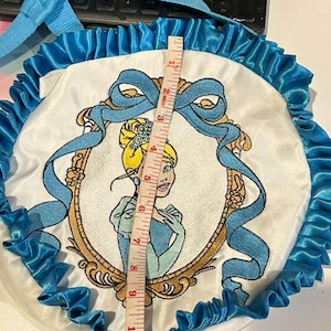 Op de afbeelding: Een ronde, witte tas met een Cinderella-thema met een blauw lint en ruches. De tas heeft een geborduurde afbeelding van Cinderella in een gouden lijst. De tas heeft een diameter van ongeveer 33 cm.