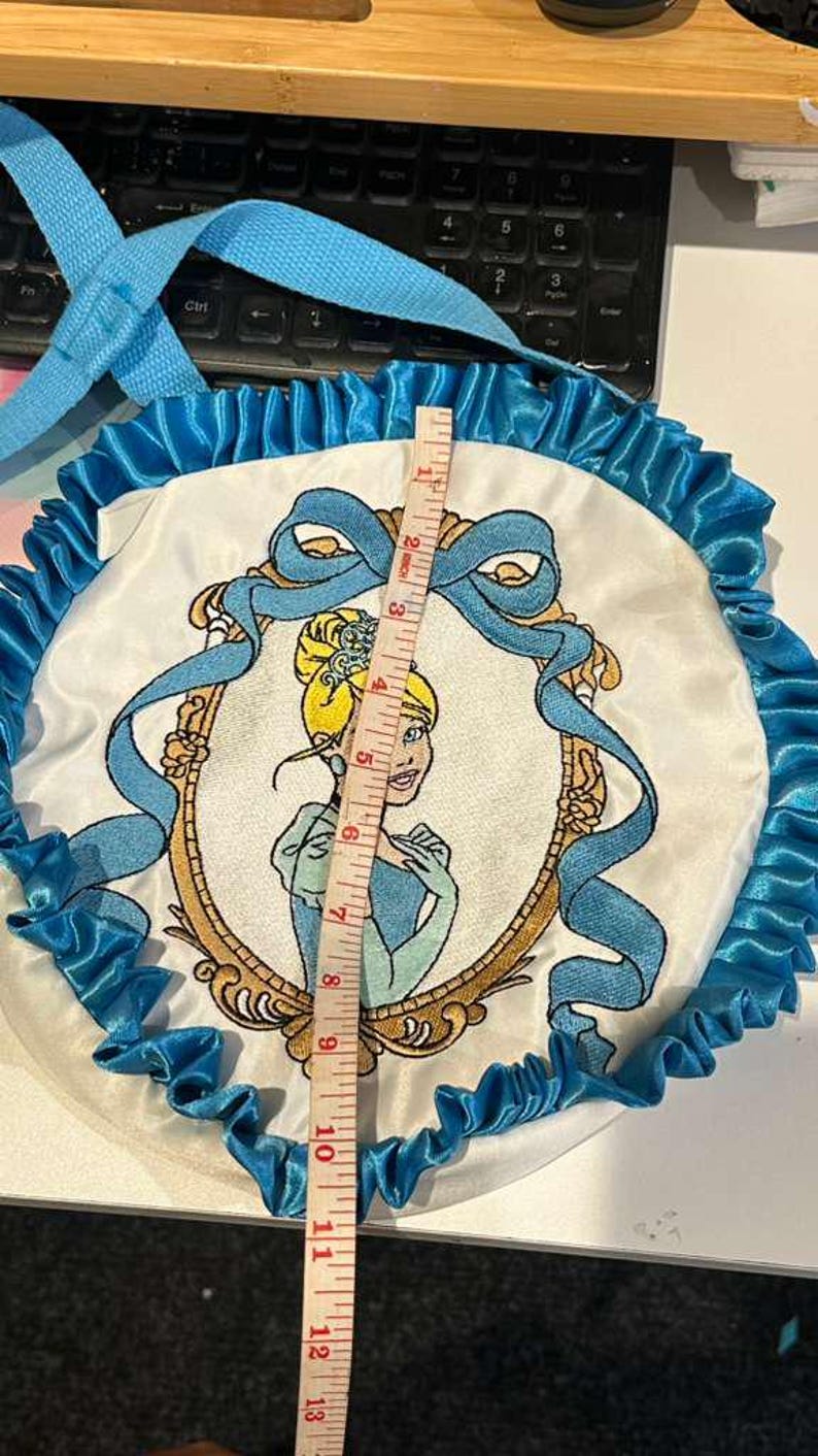 Op de afbeelding: Een ronde, witte tas met een Cinderella-thema met een blauw lint en ruches. De tas heeft een geborduurde afbeelding van Cinderella in een gouden lijst. De tas heeft een diameter van ongeveer 33 cm.