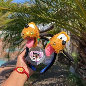 May include: Two Pluto-themed headbands with orange glitter ears and faces, black ears, and a pink tongue. The headbands are held up against a backdrop of palm leaves and a blue sky. The text "RnRcreations" is visible.