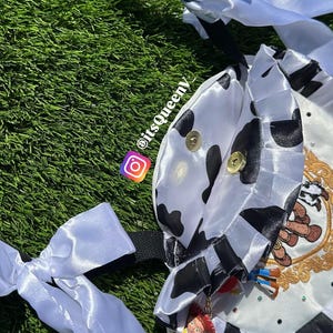 May include: A white satin bag featuring a black cow print pattern, ruffled edges, and long white ribbon ties. The bag is adorned with a gold-toned circular emblem, small rhinestones, and a dangling Jessie character charm. Text reads itsqueeny_.