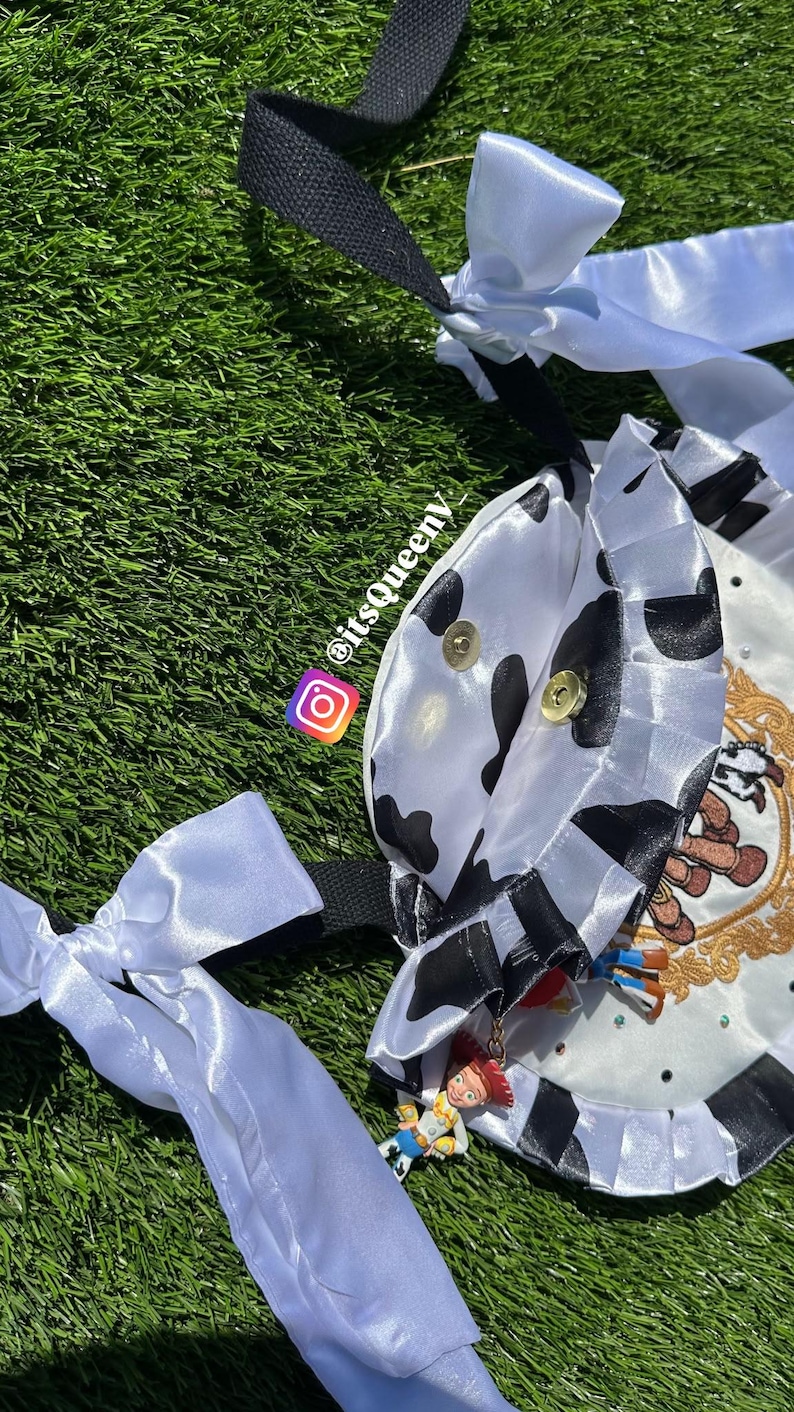 May include: A white satin bag featuring a black cow print pattern, ruffled edges, and long white ribbon ties. The bag is adorned with a gold-toned circular emblem, small rhinestones, and a dangling Jessie character charm. Text reads itsqueeny_.
