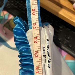 Op de afbeelding: Een blauw en wit stoffen item met een blauwe band, gemeten met een houten liniaal. De tekst op het item luidt "All around Strap size 51". De liniaal toont een meting van 129.5 cm.