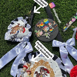 May include: A black strap with two white satin bows and two circular fabric badges featuring Toy Story character embroidery, gold-tone frames, and rhinestones. The badges have text reading Perfect to attach Any keychain and @itsQueenv.