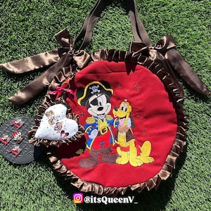 May include: Red tote bag with a brown ruffled trim and handles. The bag features an embroidered image of Mickey Mouse as a pirate with Pluto. A heart-shaped charm and pins are also visible.