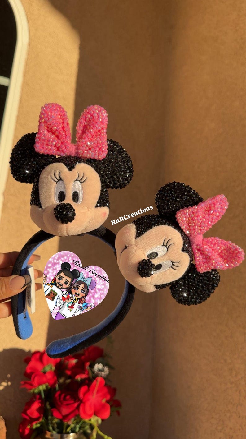 May include: Two Minnie Mouse ear headbands. One headband is held by a hand. The ears are black with pink bows. The Minnie Mouse faces are beige with black noses and eyelashes. The headband is black with blue accents. The image has the text "RnR Creations".