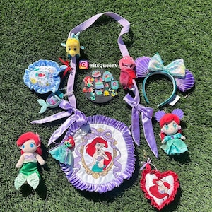 Op de afbeelding: Een verzameling Little Mermaid-thema merchandise, waaronder pluche poppen, een hoofdband met schelpoortjes, een ronde tas met een Ariel-portret en diverse accessoires. De items zijn gerangschikt op een groene ondergrond.