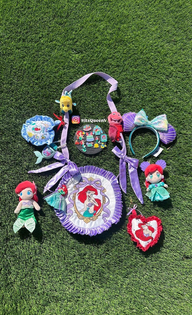 Op de afbeelding: Een verzameling Little Mermaid-thema merchandise, waaronder pluche poppen, een hoofdband met schelpoortjes, een ronde tas met een Ariel-portret en diverse accessoires. De items zijn gerangschikt op een groene ondergrond.