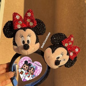 May include: Two Minnie Mouse ear headbands. The ears are black with sparkling accents, and the bows are red with silver embellishments. The headbands are black with blue accents. A heart-shaped sticker with a cartoon couple is visible.