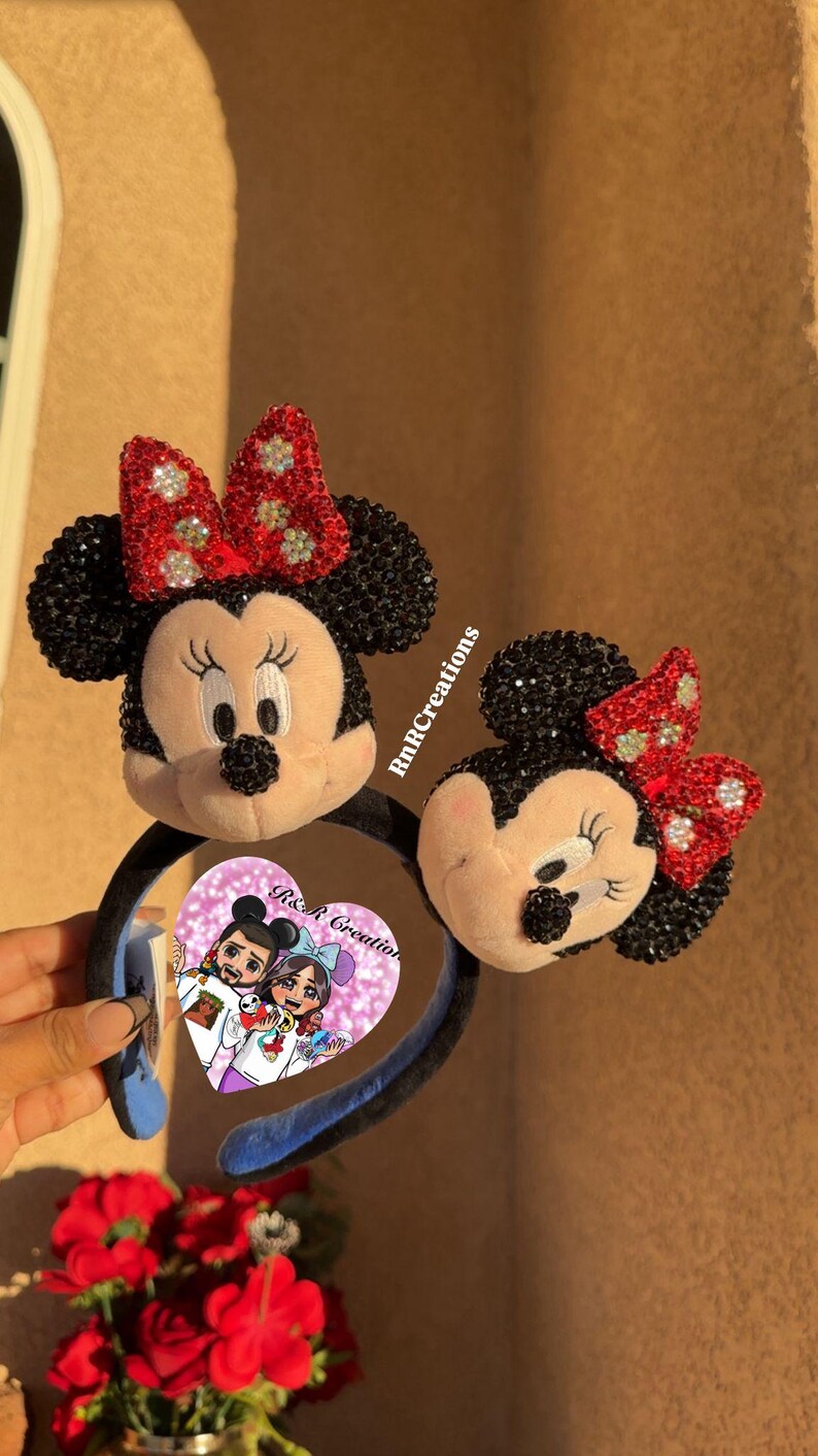 May include: Two Minnie Mouse ear headbands. The ears are black with sparkling accents, and the bows are red with silver embellishments. The headbands are black with blue accents. A heart-shaped sticker with a cartoon couple is visible.