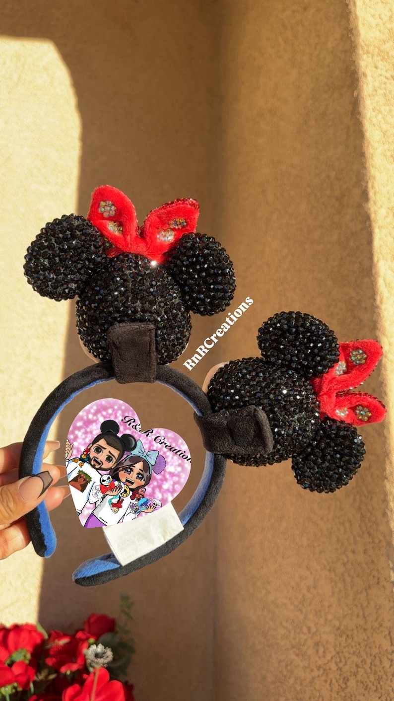 May include: Black Minnie Mouse ear headbands with red bows. The ears are covered in black rhinestones. The headband has a blue interior and a black velvet band. A heart-shaped image with cartoon characters is attached.