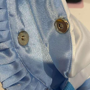 Op de afbeelding: Close-up van een lichtblauwe satijnen stof met geplooide details en een magnetische drukknoopsluiting. De stof heeft een lichte glans en de randen zijn afgewerkt met witte stof. De tas lijkt een tote of handtas te zijn.