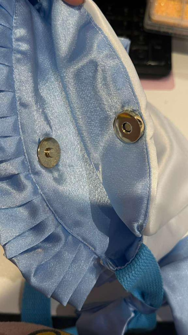 Op de afbeelding: Close-up van een lichtblauwe satijnen stof met geplooide details en een magnetische drukknoopsluiting. De stof heeft een lichte glans en de randen zijn afgewerkt met witte stof. De tas lijkt een tote of handtas te zijn.
