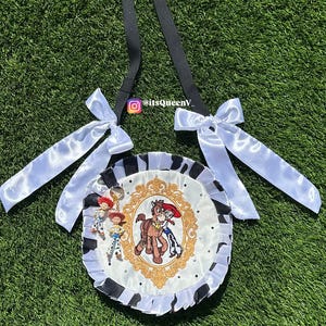 May include: A round crossbody bag featuring a white center panel with a gold-framed illustration of a cowgirl and a horse. The bag has a black and white cow-print ruffled border, two white satin bows on the black strap, and two small character charms.