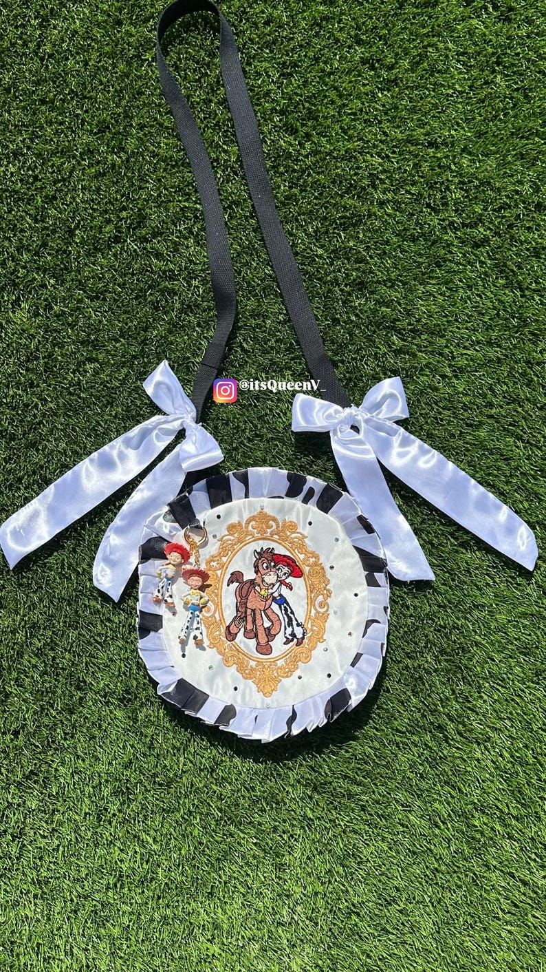 May include: A round crossbody bag featuring a white center panel with a gold-framed illustration of a cowgirl and a horse. The bag has a black and white cow-print ruffled border, two white satin bows on the black strap, and two small character charms.
